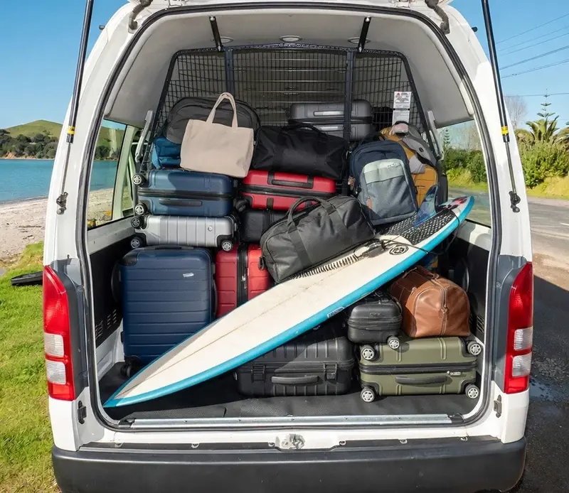 Toyota Hiace 12-seater van real photos showcasing the interior and boot space. Left: High-roof cabin with 11 grey passenger seats. Right: Open boot at Auckland Airport Terminal 3 loaded with 10+ suitcases, golf bags (Callaway and Titleist), stroller and luggage.